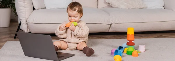 Toddler girl holding toy and looking at laptop at home, banner — Stock Photo