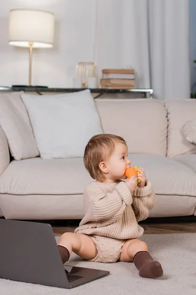 Vista lateral de la niña sosteniendo juguete cerca de la computadora portátil en casa en la sala de estar - foto de stock