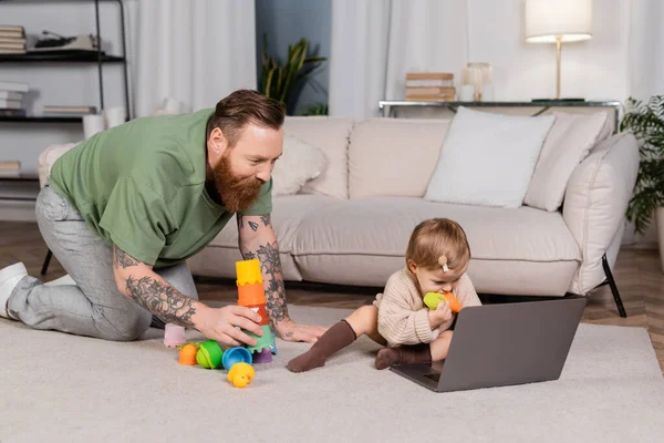 Tattooed father holding toys near baby daughter and laptop at home - foto de stock