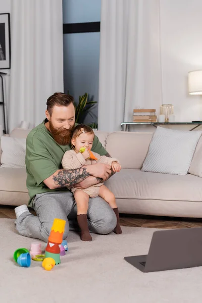 Papá barbudo sosteniendo a su hija con juguete cerca de la computadora portátil en la sala de estar - foto de stock