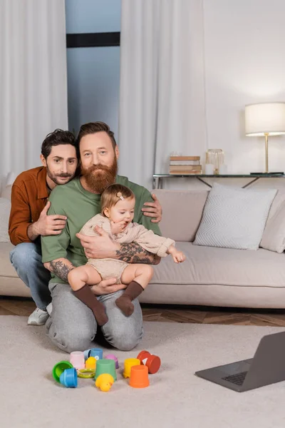 Same sex couple with toddler daughter looking at camera near toys and laptop in living room - foto de stock