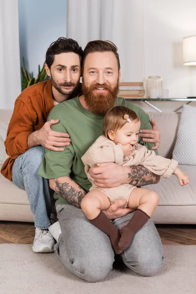 Gay couple holding baby daughter and looking at camera in living room - foto de stock