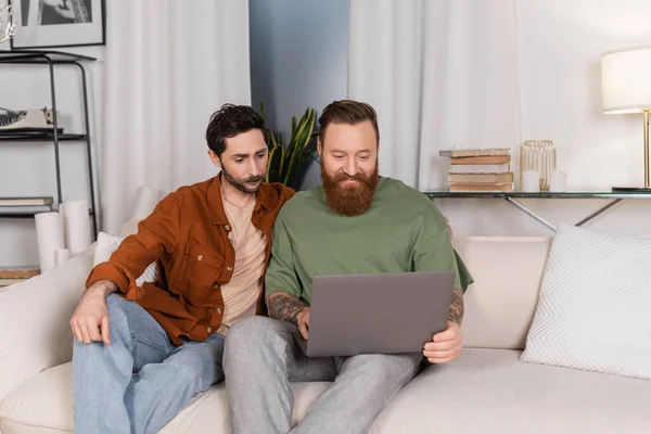 Sonriente gay hombre usando laptop cerca pareja en casa - foto de stock