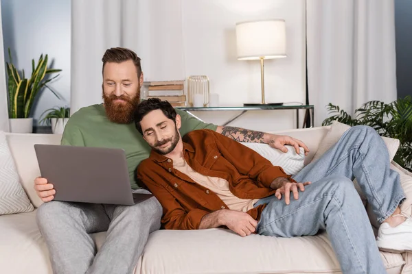 Positive same sex couple using laptop on couch at home - foto de stock