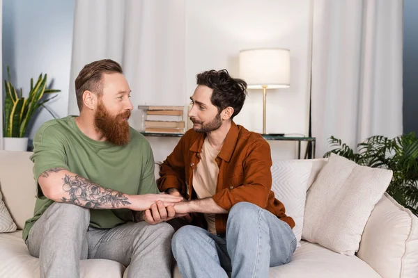 Barbudo gay pareja cogida de la mano en sofá en casa - foto de stock