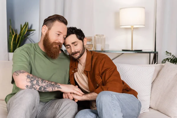 Gay man holding hand of partner while sitting on couch at home - foto de stock