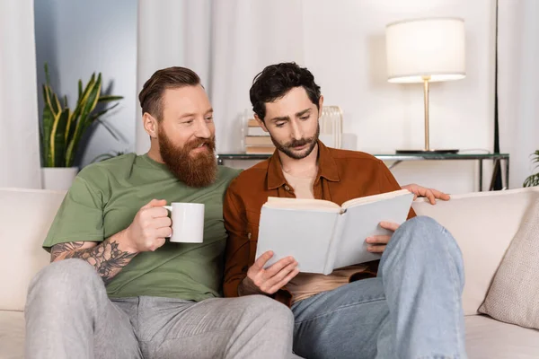 Усміхнений гей чоловік тримає чашку кави, коли партнер читає книгу вдома — стокове фото