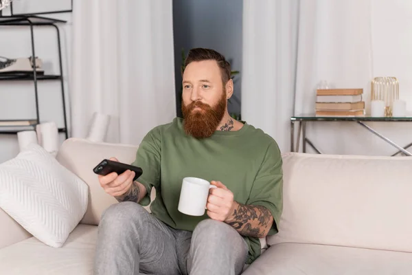 Татуированный мужчина держит чашку кофе и смотрит телевизор на диване дома — стоковое фото