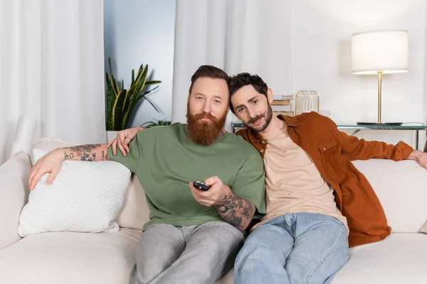 Bearded same sex couple watching tv on couch at home - foto de stock