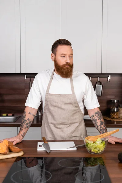 Tattooed man in apron standing near fresh salad in kitchen — стоковое фото