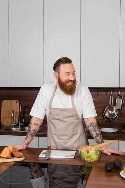 Cheerful tattooed man in apron standing near food and fresh salad in kitchen — Stock Photo