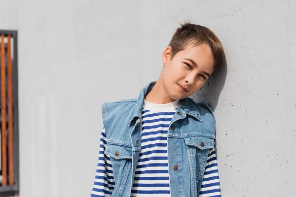 Niño preadolescente bien vestido en chaleco vaquero y camisa de manga larga a rayas apoyada en la pared gris - foto de stock