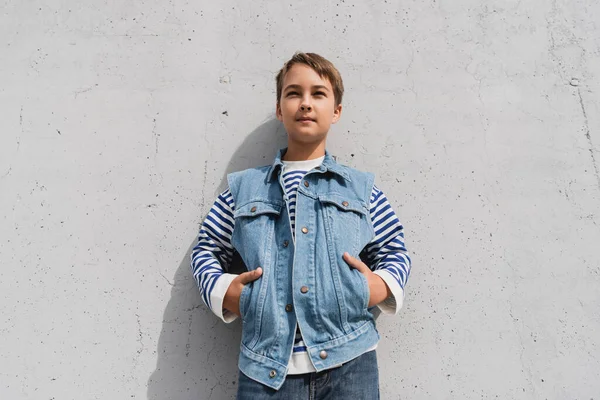 Stylish preteen boy in denim vest and striped long sleeve shirt posing with hands in pockets near mall with grey wall — Photo de stock