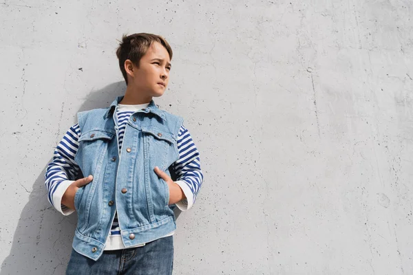 Low angle view of stylish boy in denim vest and striped long sleeve shirt posing with hands in pockets near mall with grey wall — Photo de stock