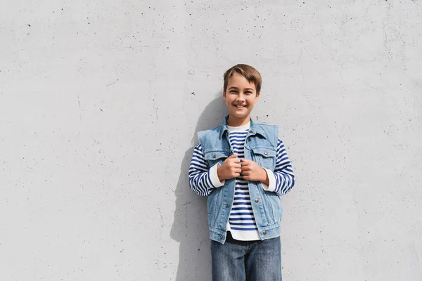 Happy boy in denim vest and striped long sleeve shirt standing near mall with grey wall — Photo de stock