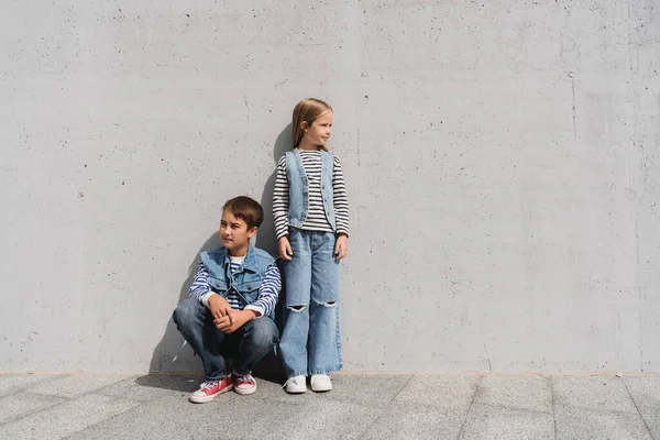 In voller Länge stylische Kinder in Jeans-Outfits mit gestreiften Langarmhemden, die in der Nähe der grauen Wand in einem Einkaufszentrum posieren — Stockfoto