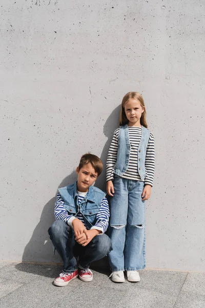 Full length of stylish preteen kids in denim outfits posing near grey wall in mall — Stock Photo