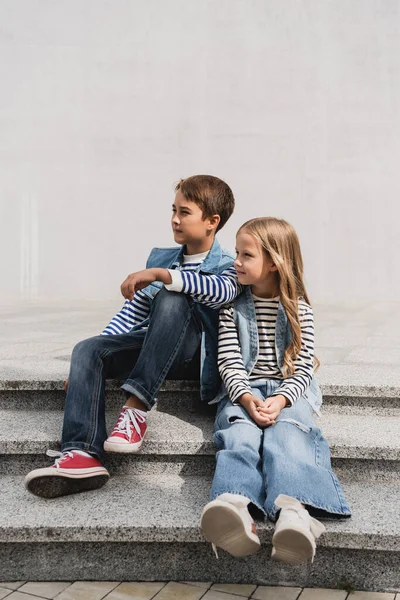 In voller Länge gut gekleidete Kinder in Jeans-Outfits sitzen auf Treppen in der Nähe des Gebäudes — Stockfoto