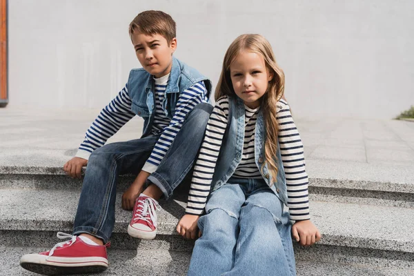 Well dressed children in denim vests with long sleeve shirts sitting on stairs near mall — стоковое фото