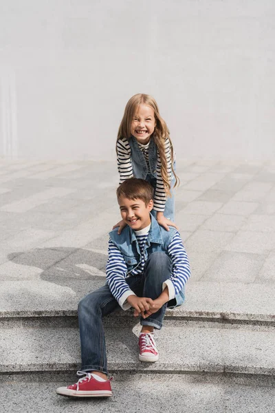 Cheerful girl in stylish clothes leaning on shoulders of happy preteen boy sitting on stairs — Fotografia de Stock