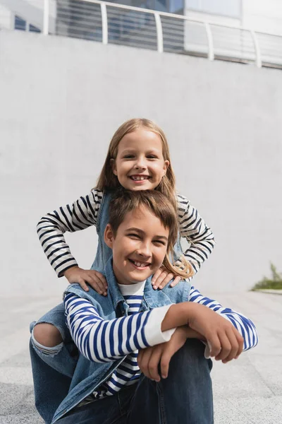 Pleased girl in stylish clothes leaning on shoulders of happy well dressed boy near mall — Stock Photo