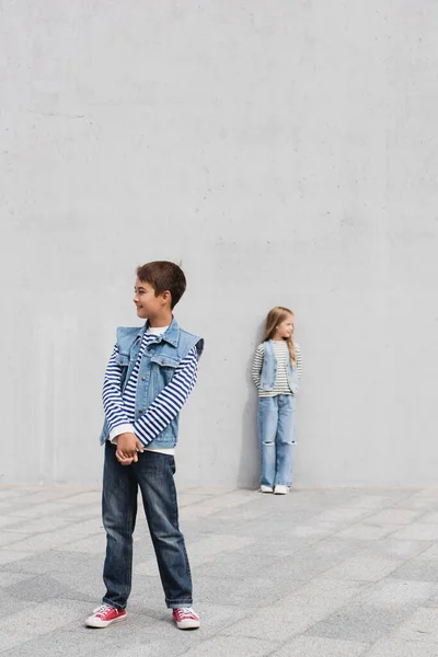 Full length of cheerful boy in stylish denim outfit standing near girl on blurred background - foto de stock