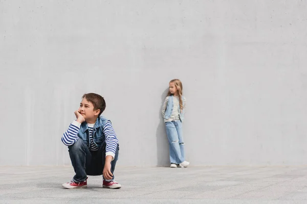 Full length of stylish boy in denim outfit sitting near girl on blurred background - foto de stock