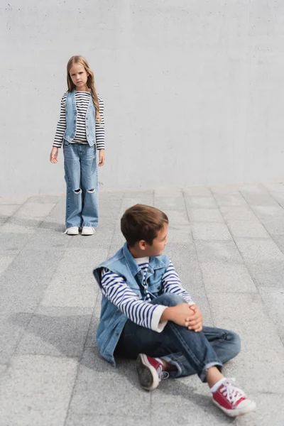 Comprimento total de menina bem vestida em roupa de ganga em pé perto do menino em primeiro plano desfocado — Fotografia de Stock