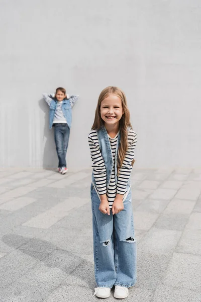 Повна довжина веселої дівчини в модному джинсовому вбранні, що стоїть біля хлопчика на розмитому фоні — стокове фото