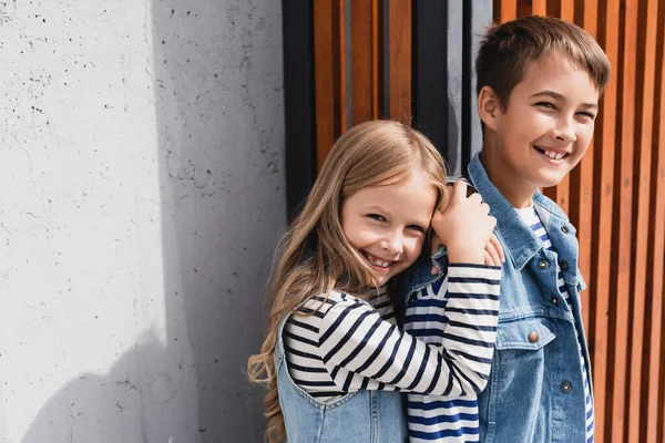 Porträt fröhlicher Kinder in gestreiften langärmeligen Hemden und Jeanswesten, die in der Nähe eines Gebäudes in die Kamera schauen — Stockfoto