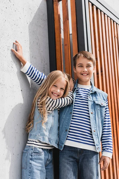 Portrait of happy children in striped long sleeve shirts and denim vests looking at camera near building — Stock Photo