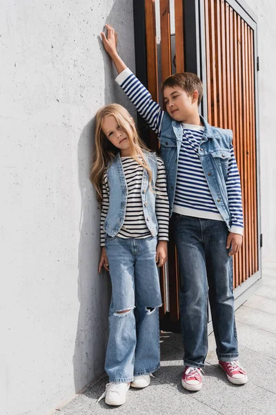 Полная длина стильных детей в полосатых футболках с длинными рукавами и джинсовых жилетах, позирующих рядом со зданием — стоковое фото
