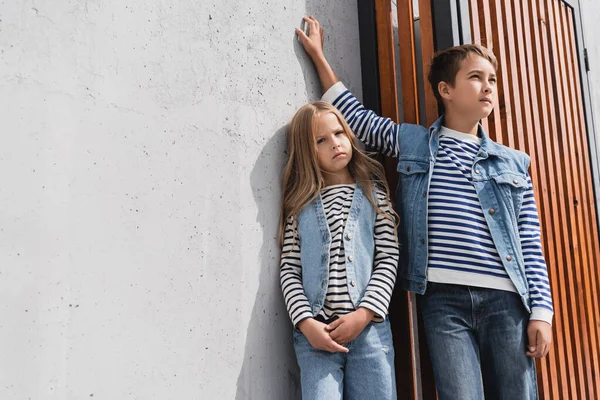 Stylish preteen boy in denim vest leaning on wall while standing with girl near mall - foto de stock