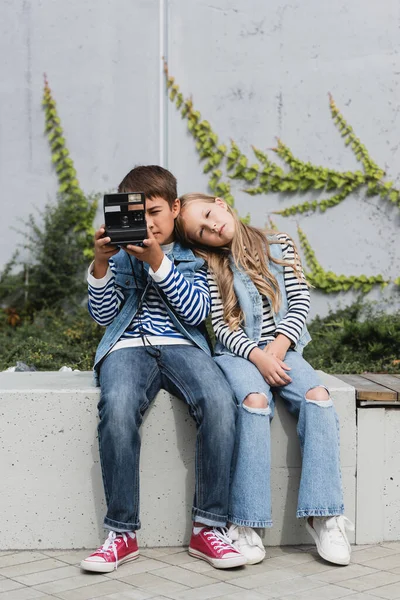 Niño preadolescente en ropa elegante tomando fotos en la cámara vintage cerca de la chica en chaleco vaquero y jeans - foto de stock