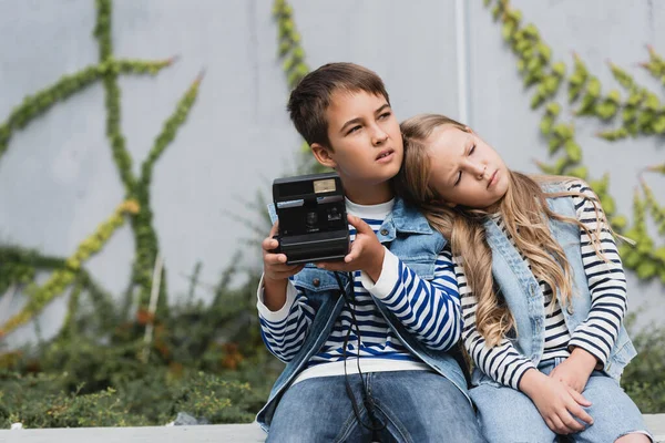 Girl in denim vest and jeans leaning on shoulder of preteen boy in stylish clothes holding vintage camera — стоковое фото