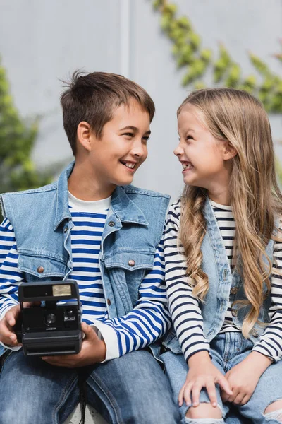 Щасливий хлопчик у стильному одязі тримає старовинну камеру біля добре одягненої дівчини — стокове фото