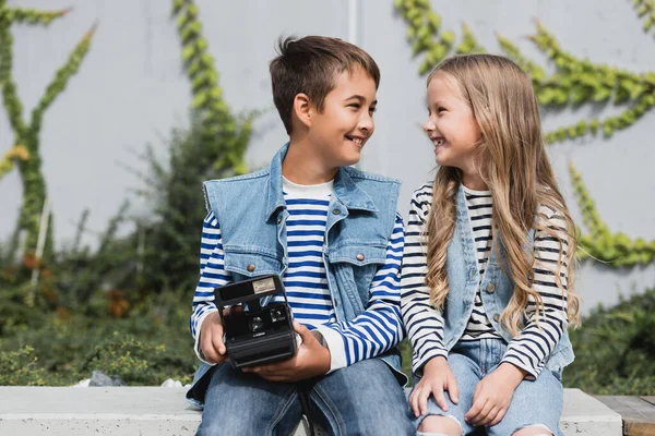 Happy preteen boy in stylish clothes holding vintage camera near smiling girl — Fotografia de Stock