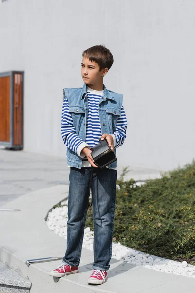 Full length of well dressed preteen boy in denim clothes holding vintage camera outside - foto de stock