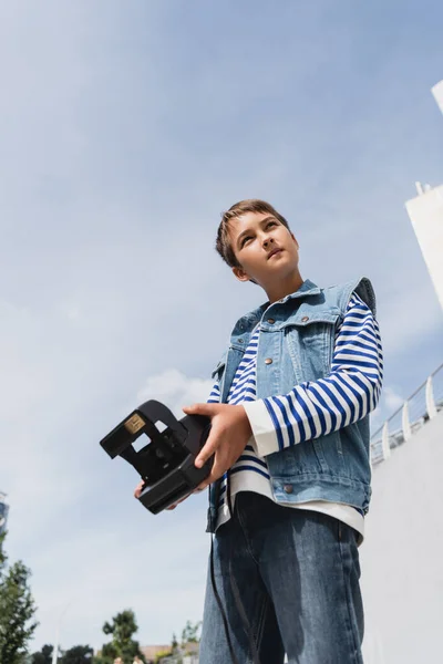 Vue à angle bas de garçon préadolescent bien habillé en tenue denim tenant caméra vintage à l'extérieur — Photo de stock