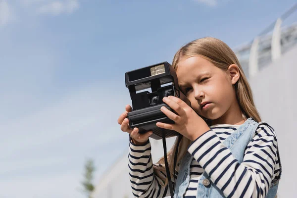 Preteen girl in denim vest and striped long sleeve shirt taking photo of vintage camera — Stock Photo