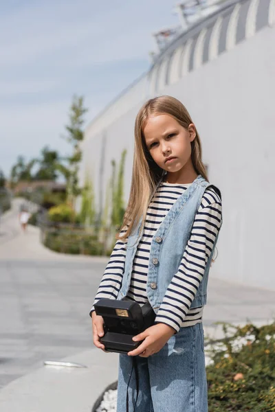 Preteen girl in denim vest and striped long sleeve shirt holding vintage camera near mall building — Stock Photo