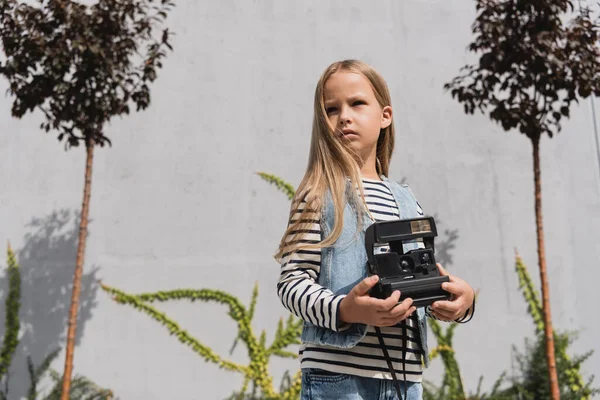 Girl in blue denim vest and striped long sleeve shirt holding vintage camera near mall building — Stock Photo