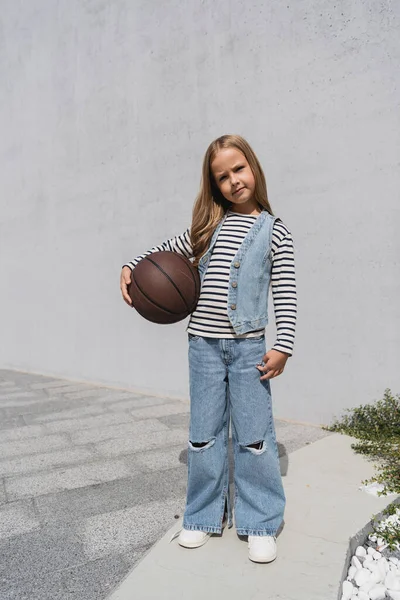 Pleine longueur de fille préadolescente en gilet en denim et jeans bleus tenant basket près du bâtiment du centre commercial — Photo de stock