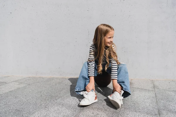 Comprimento total de menina sorridente em colete jeans jeans jeans jeans e azul sentado no basquete perto da parede — Fotografia de Stock