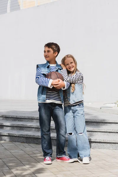 Preteen boy in denim vest holding basketball near happy girl while standing near mall — Fotografia de Stock