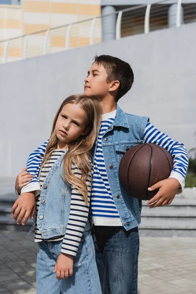 Мальчик в джинсовом жилете держит баскетбол и обнимает стильную девушку, стоя возле торгового центра — стоковое фото