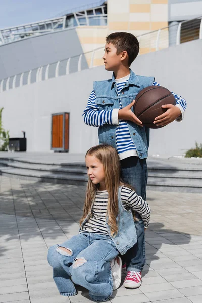 Menina em roupas elegantes abraçando as pernas do menino com basquete em pé perto do shopping — Fotografia de Stock
