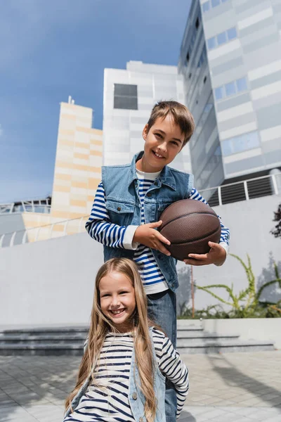 Allegro ragazzo in gilet di jeans che tiene il basket vicino sorridente ragazza mentre in piedi vicino al centro commerciale — Foto stock