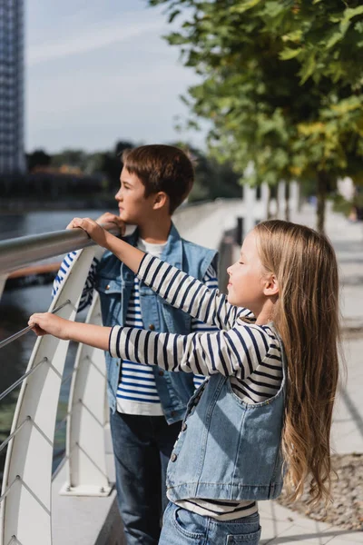 Stylish kids in denim vests standing near metallic fence on riverside — стоковое фото