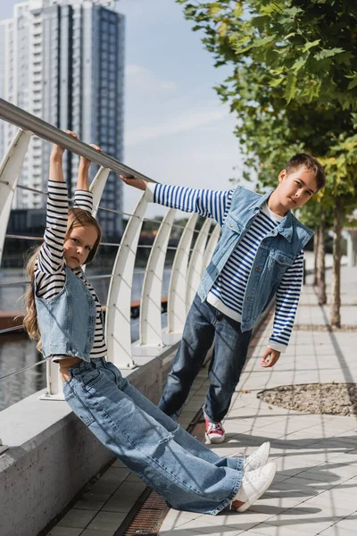 Повна довжина стильних дітей в джинсових жилетах і джинсах, що позують біля металевого паркану на березі річки — стокове фото